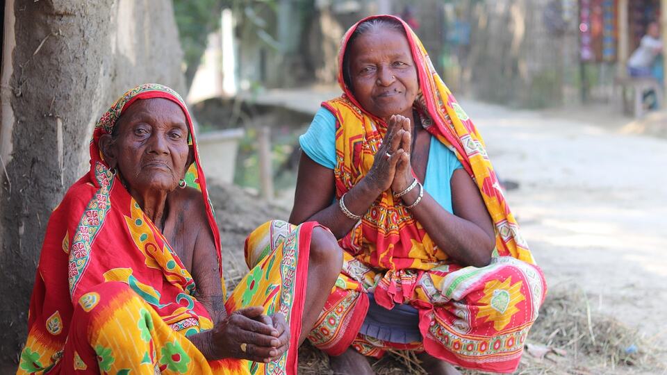 Two Indian women in colourful saris smiling, one greeting with folded hands